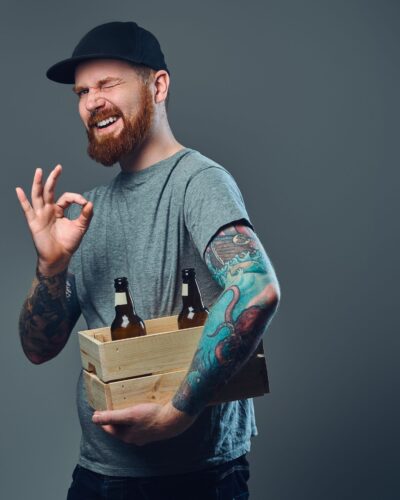 Portrait of bearded male in a cap with tattoos on his arms holds a wooden box with beer bottles.